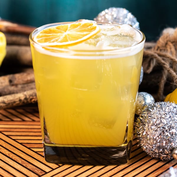 A yellow cocktail with ice and a dried lemon slice, surrounded by festive silver ornaments.