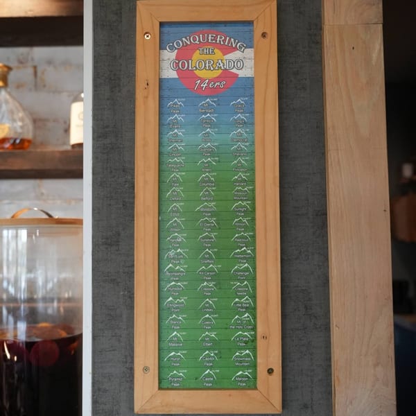 A wooden framed sign with a Colorado flag background lists "Conquering the Colorado 14ers" and peak names.