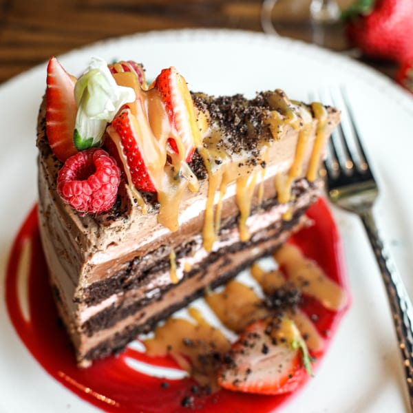 A slice of chocolate cake with caramel drizzle, strawberries, raspberries, and cookie crumbles.