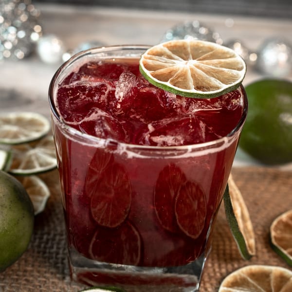 A vibrant red cocktail with ice and a dried lime slice, surrounded by limes and festive decor.