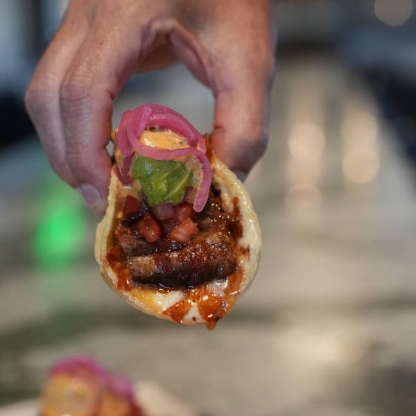 Hand holding a West Main Taproom taco with meat, salsa, guacamole, and pickled onions.
