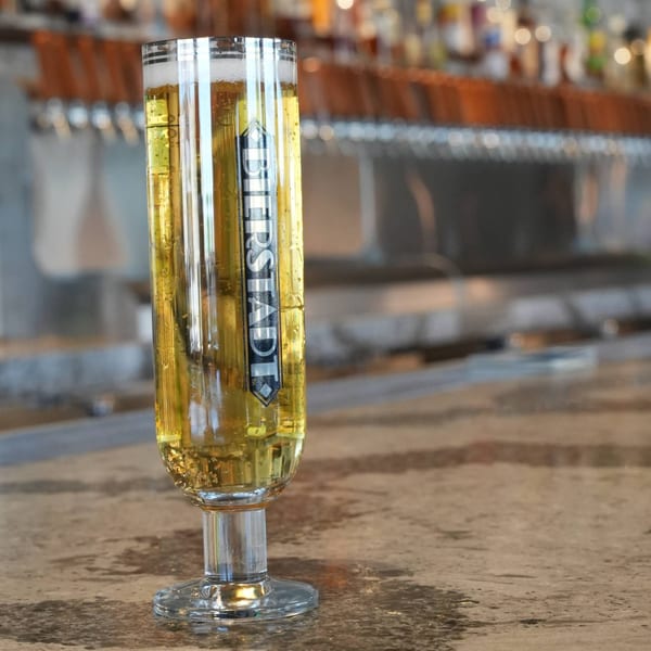 A tall glass of golden beer with a Bierstadt logo sits on a bar counter with taps in the background.