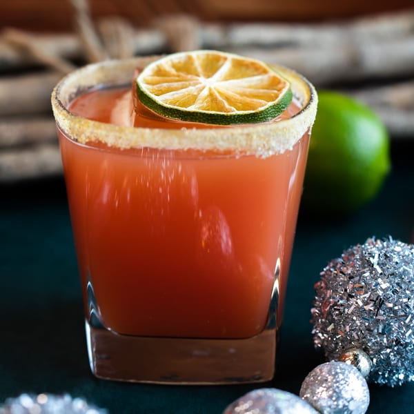 A red cocktail with a sugar-salted rim and a dried lime slice, next to a fresh lime and silver ornaments.