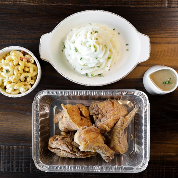 Fried chicken in a foil pan, mashed potatoes, macaroni and cheese, and gravy on a wooden table.