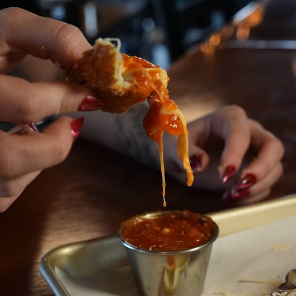 A hand holds a mozzarella stick with melted cheese and sauce dripping into a small metal cup.