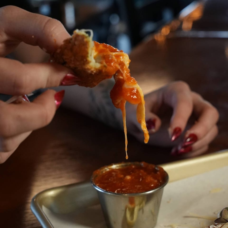 A hand holds a mozzarella stick with melted cheese and sauce dripping into a small metal cup.