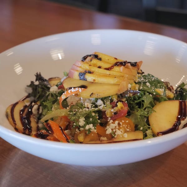 A vibrant salad in a white bowl, featuring greens, sliced peaches, crumbled cheese, and balsamic glaze.