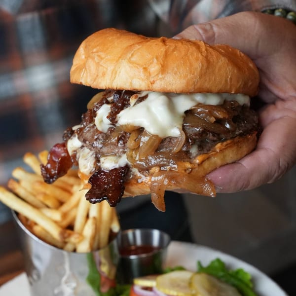 A hand holds a West Main Taproom burger with caramelized onions, bacon, and melted cheese.