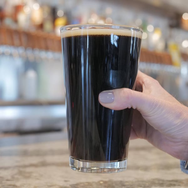 A hand holds a pint glass of dark beer with a foamy head at West Main Taproom.