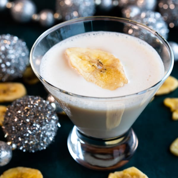 A creamy cocktail with a banana chip garnish, surrounded by silver ornaments and dried banana slices.