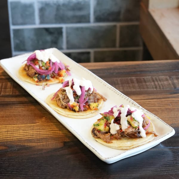 Three tacos with shredded meat, pineapple salsa, pickled red onions, and crema on a white platter.