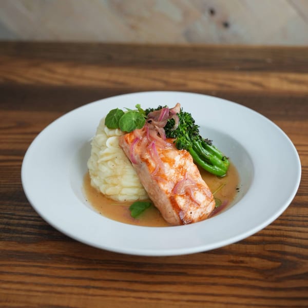 Salmon fillet with pickled red onions, mashed potatoes, broccolini, and a light sauce on a white plate.